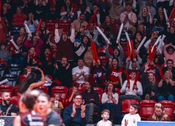 A Crowd of 'The Tribe' cheering wearing 'The Tribe' T-shirts holding long inflatables colours red and white