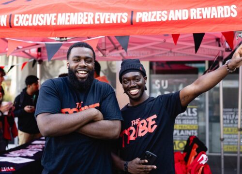 Two Students Wearing 'The Tribe' T-shirts in front of a shall